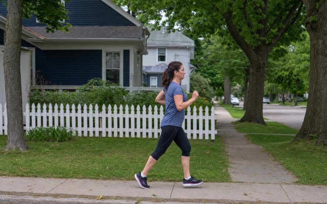 A woman running with relaxed confidence along a tree-lined residential sidewalk in daylight, seen from the side