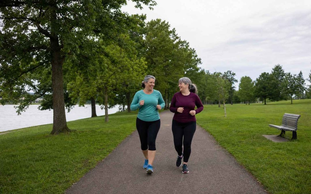 Two women in their early fifties jogging side by side along a tree-lined park trail